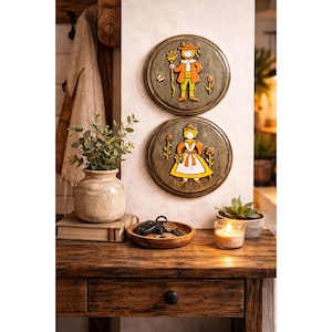 May include: Two round, wooden wall plaques with folk art figures of a man and woman in traditional attire. The plaques are green with yellow, orange, and white details. The scene is set on a wooden table with a vase, books, and a candle.