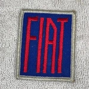 May include: A rectangular patch featuring the word "FIAT" in red, embroidered on a blue background. The patch has a silver border and is likely a logo or emblem.