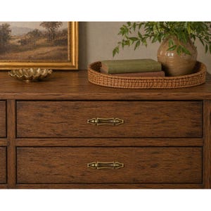 May include: A wooden dresser with two drawers, featuring brass hardware. A woven tray holds books and a vase with greenery. A gold-colored decorative bowl and a framed landscape painting are also visible.