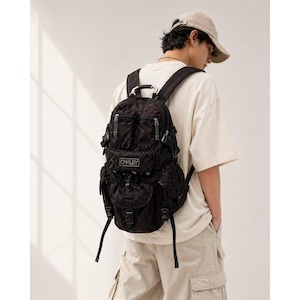 May include: Black Oakley backpack with multiple compartments and straps. The backpack has the Oakley logo on the front. The backpack is worn by a person wearing a beige t-shirt and khaki pants.