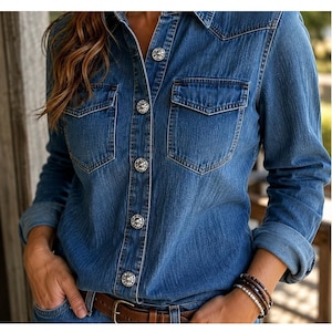 May include: A long-sleeved blue denim shirt featuring two chest pockets with flaps and a front button-down closure. The shirt is secured with round, silver-toned metal buttons that have an embossed decorative pattern on their surface.
