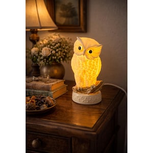 Puede incluir: Lámpara con forma de búho de cerámica que emite una cálida luz amarilla. La lámpara tiene texturas de plumas detalladas y se asienta sobre una base decorativa. La lámpara de búho está sobre una mesita de noche de madera.
