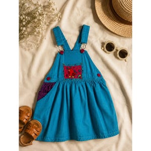 Puede incluir: Un vestido peto de mezclilla azul con un diseño de bolsillo colorido. El vestido tiene botones rojos y forro de bolsillo morado. También hay sandalias de cuero marrón y gafas de sol en la imagen. El vestido es para niños.