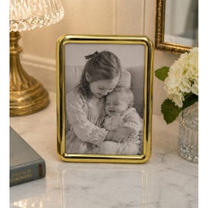 May include: A gold-colored rectangular picture frame with rounded corners. The frame holds a black and white photograph of two children. The frame is displayed on a marble surface, next to a book and a lamp.