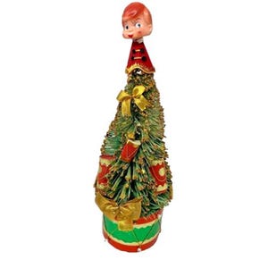 May include: A vintage Christmas tree decoration featuring a boy's head atop a green tree. The tree is adorned with gold bows, red drums, and a red and gold drum base. The boy has red hair and is wearing a red hat.