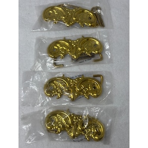 Op de afbeelding: Vier nieuwe, goudkleurige gespen met sierlijke, symmetrische ontwerpen. Elke gesp is verpakt in een doorzichtige plastic zak. De gespen hebben een klassieke, vintage uitstraling, met een haak- en lus sluiting.