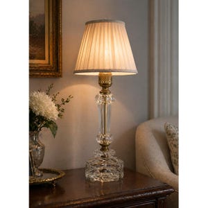 May include: A crystal table lamp with a pleated lampshade. The lamp has a clear, faceted base and a brass-colored neck. The lampshade is a light beige color. The lamp is on a dark wooden table.