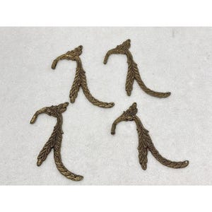 May include: Four antique bronze-colored decorative metal hooks. Each hook has a curved shape with a leaf-like design. The hooks are arranged on a white surface, showcasing their intricate details and vintage aesthetic.