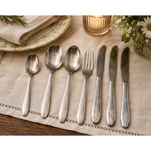 May include: A set of seven silver-toned metal utensils arranged in a row, including one small spoon, two large spoons, one fork, and three knives. Each piece features a smooth, polished finish with a subtle engraved detail on the handle.