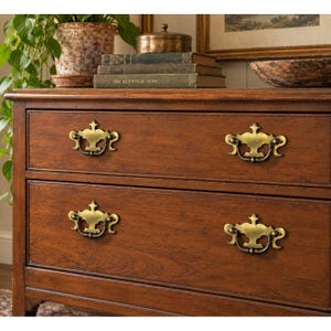May include: A wooden chest of drawers with a warm brown finish, featuring two drawers each equipped with two ornate brass-toned metal swing handles. The top surface holds a stack of three books, including one titled The Kinfolk Home.