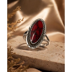 Puede incluir: Un anillo plateado con una piedra roja ovalada. La piedra está engastada en un bisel decorativo con un diseño de cuerda trenzada. El anillo descansa sobre una superficie texturizada de color marrón claro.