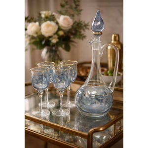 May include: A clear glass decanter with a blue floral design and a matching stopper, alongside four wine glasses with gold rims and blue floral patterns. The set is displayed on a gold-rimmed tray, suggesting a bar or dining setting.