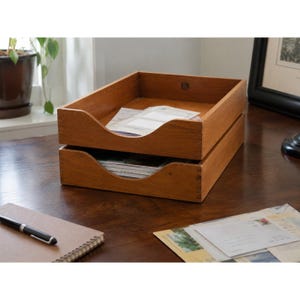 May include: A stack of two rectangular wooden document trays with a medium brown finish and visible joinery details. The front panels feature a curved cutout for easy access to papers, with white documents resting inside both of the trays.