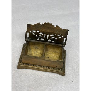 May include: A vintage brass box with a hinged lid and two compartments. The lid features a decorative, openwork design. The box sits on a rectangular base with a beaded border. The overall color is a warm, aged brass.