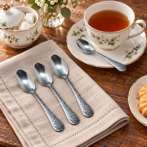 Puede incluir: Cuatro cucharas de metal plateado con un acabado martillado y texturizado en los mangos. Tres cucharas están dispuestas en diagonal sobre una servilleta de lino beige, mientras que una cuchara descansa sobre un plato de porcelana blanca.
