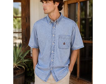 Puede incluir: Una camisa de mezclilla azul claro de manga corta con cuello clásico, tapeta de botones frontal y un bolsillo en el pecho en el lado izquierdo. El bolsillo muestra un pequeño logotipo circular bordado en tonos marrones y rojos.
