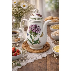 Può includere: Caffettiera in ceramica bianca con un motivo floreale con un fiore viola e farfalle. La caffettiera è appoggiata su un sottobicchiere intrecciato, con una tazza di caffè, fragole e zollette di zucchero nelle vicinanze. Un mazzo di fiori bianchi è sullo sfondo.