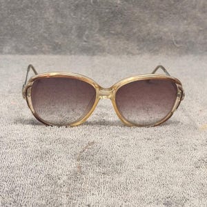 May include: Sunglasses with a translucent, light amber-colored plastic frame and gradient-tinted lenses. The frame features a rounded, oversized shape with thin, metallic-toned temples extending from the sides, providing a clear view of the front.