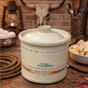 Puede incluir: Una olla de cocción lenta Rival Little Dipper de color crema con tapa. La olla de cocción lenta tiene una banda decorativa con un diseño del suroeste. El fondo incluye una linterna, una cuerda y una calavera de toro.