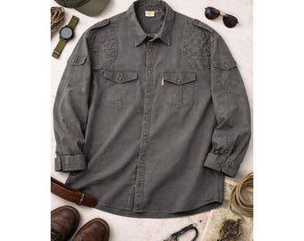 Puede incluir: Una camisa gris de manga larga con dos bolsillos en el pecho y parches en los hombros. La camisa es abotonada y tiene cuello. Los accesorios incluyen una gorra verde, reloj, gafas de sol, cinturón, botas y una brújula.