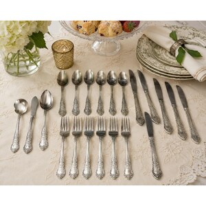 May include: A set of silver-toned flatware featuring ornate floral embossed handles, including various spoons, forks, and knives arranged on a cream-colored tablecloth alongside a glass vase of white flowers and a stack of patterned plates.