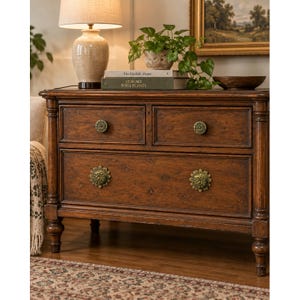May include: A dark brown wooden chest of drawers featuring two small top drawers and one large bottom drawer, all equipped with ornate, circular brass-toned knobs. The piece stands on four turned legs and displays a rich, polished wood grain finish.