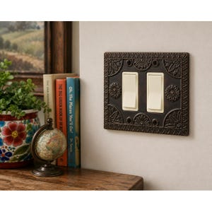 May include: A double light switch wall plate featuring an ornate, dark bronze-toned metal finish with embossed floral and leaf patterns. The rectangular plate includes two cream-colored rocker switches centered within the decorative frame.