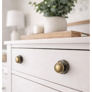 May include: Close-up of a white dresser with two decorative brass knobs. The knobs have a detailed design with a circular center and a textured outer ring. A wooden tray with a plant and candle sits on top.