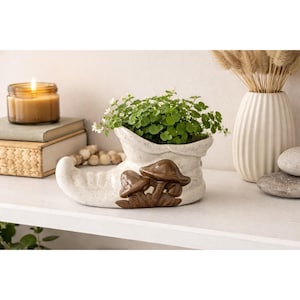 May include: A ceramic planter shaped like an elf boot, filled with green plants and small white flowers. The boot is off-white with brown mushroom accents. A lit candle, books, and a vase with dried grass are in the background.