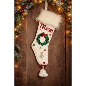 Puede incluir: Calcetín navideño color crema con la palabra "Mom" en escritura roja. Presenta una corona verde con un lazo rojo, lentejuelas y un puño de piel sintética blanca. Una pequeña campana de ganchillo cuelga de la parte inferior.