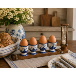 May include: A wooden stand holding four blue and white ceramic egg cups, each containing a brown egg, and a small wooden hourglass. The egg cups feature a windmill pattern. The stand is rectangular with metal accents at the corners.