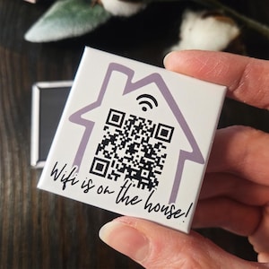 May include: A white square magnet with a purple house outline, a QR code, and a WiFi symbol. The text "Wifi is on the house!" is written below the QR code. The magnet is held in a person's hand.