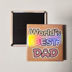 May include: A square magnet with the text "World's BEST DAD" in a playful font. The magnet has a light brown background with a holographic effect. The text is outlined in black with pink and blue accents. A second magnet is visible.