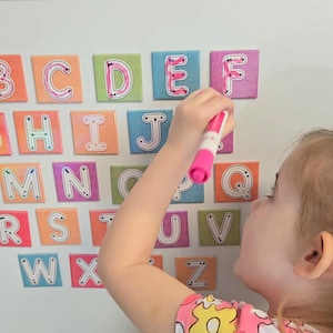 May include: Colorful alphabet magnets arranged on a white surface. Each magnet features a letter outlined in a contrasting color against a square background. A pink marker is held near the letters, suggesting educational play.