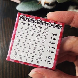 May include: A small, rectangular kitchen conversion chart with a red and white checkered border. The chart lists conversions for cups, ounces, tablespoons, teaspoons, and milliliters, along with equivalent measurements for gallons, quarts, pints, and cups.