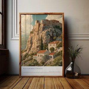 Może przedstawiać: Oprawiony wydruk z twierdzy Klis w Chorwacji, przedstawiający szczegółową ilustrację zabytkowego miejsca. Grafika przedstawia twierdzę na skalistej górze, z budynkami i drzewami. Wydruk jest w drewnianej ramie, eksponowany na drewnianej podłodze.