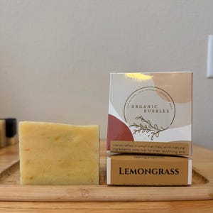 Könnte beinhalten: Ein Stück handgemachte Lemongrass-Seife neben einer Schachtel mit dem Text "Organic Bubbles" und "Lemongrass". Die Seife ist hellgelb mit sichtbaren Flecken. Die Schachtel hat ein rotes und beiges Design.