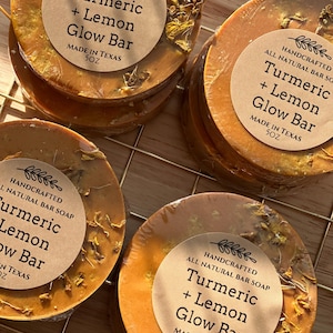 Puede incluir: Una pila de barras redondas de color amarillo anaranjado de cúrcuma y limón. Cada barra tiene una etiqueta marrón que dice "Turmeric + Lemon Glow Bar" y "Made in Texas 5OZ". Las barras están dispuestas sobre una rejilla de alambre dorada.