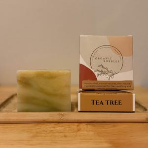 Op de afbeelding: Een vierkant stuk zeep met een gemarmerde, lichtgroene en gele uitstraling, naast twee dozen. Op de bovenste doos staat "ORGANIC BUBBLES" en op de onderste doos "TEA TREE". De zeep en dozen staan op een houten oppervlak.