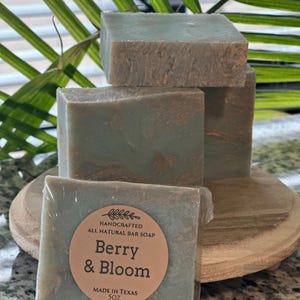Könnte beinhalten: Handgemachte Naturseife mit der Aufschrift "Berry & Bloom" auf einem Holzständer gestapelt. Die Seife ist ein gedecktes Grün mit brauner Marmorierung. Das Etikett gibt an, dass die Seife in Texas hergestellt wurde und 142 Gramm wiegt.