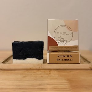 Peut inclure: Un pain de savon artisanal avec le dessus noir et le dessous blanc, à côté de deux boîtes. Les boîtes indiquent "Organic Bubbles" et "Vetiver & Patchouli". Le savon et les boîtes sont sur un plateau en bois.
