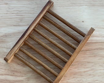 May include: A wooden soap dish with a slatted design. The dish is made of light brown wood and has a rectangular shape with parallel wooden slats. The dish is designed to allow water to drain, extending the life of the soap.