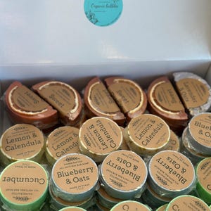 May include: A box of handcrafted natural bar soaps with various scents, including lemon & calendula, blueberry & oats, and cucumber. Each soap is round or half-circle shaped, with labels indicating the scent and origin. The box has the text "Organic bubbles".