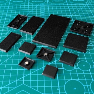 Könnte beinhalten: Eine Ansammlung schwarzer, rechteckiger Komponenten mit strukturierten Oberflächen und verschiedenen Formen. Einige Teile haben kleine, kreisförmige Vertiefungen, während andere eingebaute Magnete aufweisen. Die Artikel sind auf einer grünen Schneidematte angeordnet.