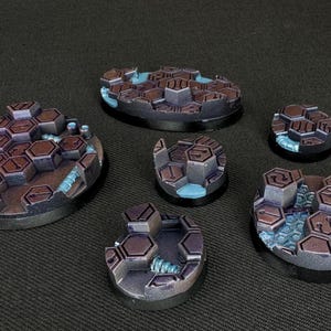 Op de afbeelding: Zes ronde bases van verschillende grootte voor miniatuurfiguren, geschilderd in een donkerbruin en blauw kleurenschema. De bases hebben een getextureerd oppervlak met een honingraatpatroon.