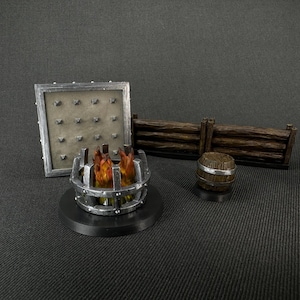 Op de afbeelding: Een miniatuur tabletop-spelset met een metalen vuurplaats, een houten hek en een metalen muur met een geniet oppervlak. De vuurplaats staat op een zwarte basis en het hek is gemaakt van bruin hout.