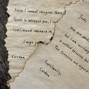 May include: Close-up of several pieces of aged paper with handwritten text in black ink. The paper edges are torn and singed, giving a vintage appearance. The text includes names like "Jude" and "Cardan" and phrases like "I cannot imagine" and "The treaties are signed."