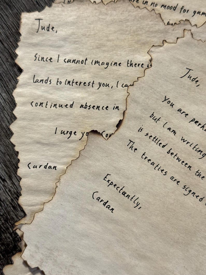 May include: Close-up of several pieces of aged paper with handwritten text in black ink. The paper edges are torn and singed, giving a vintage appearance. The text includes names like "Jude" and "Cardan" and phrases like "I cannot imagine" and "The treaties are signed."