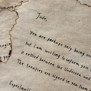 May include: Aged parchment paper with handwritten text. The letter is addressed to "Jude" and signed "Cardan." The edges of the paper are torn and singed, giving it an antique appearance. The text is in black ink.