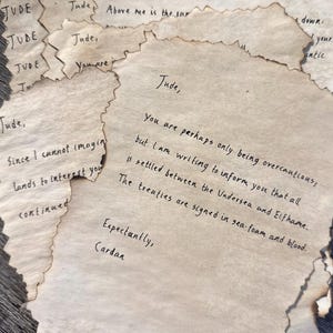 May include: A collection of aged paper notes with handwritten text. The edges of the paper are singed, giving them a vintage appearance. The text includes phrases like "To the High Queen of Elfhame" and "The treaties are signed in sea-foam and blood."
